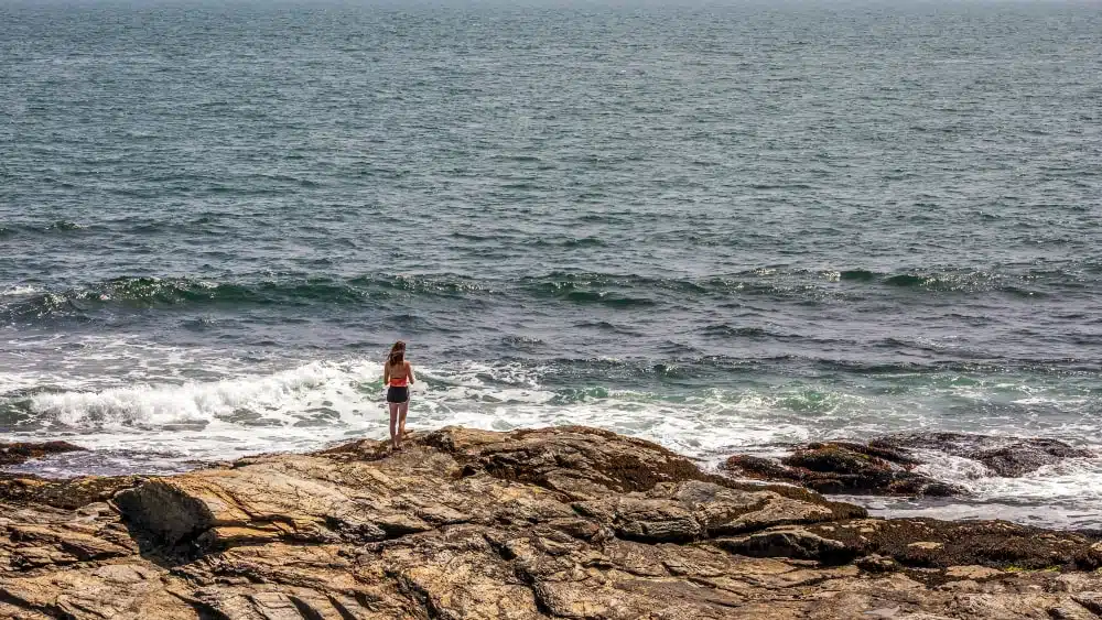 A young woman stands on rocky beach overlooking waves at Beavertail State Park, Rhode Island