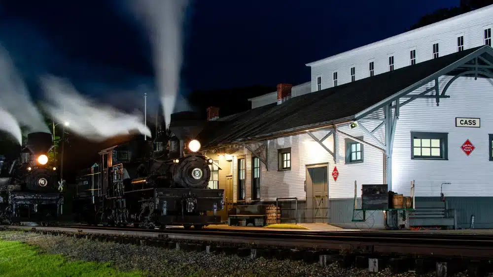 Two black locomotors outside of a historic train stat6ion with white siding and a black roof.