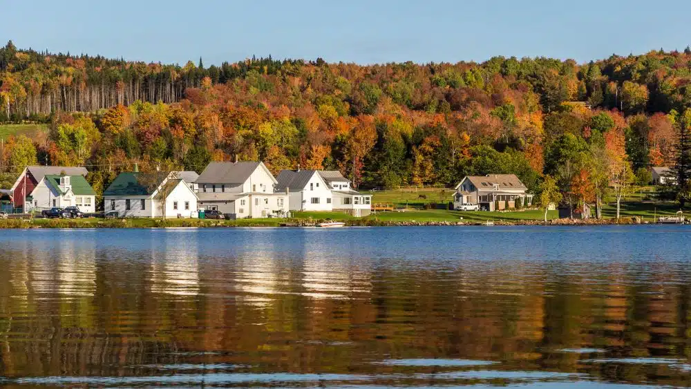 Large houses along the edge of a lake with a green and yellow and orange and red forest behind them.