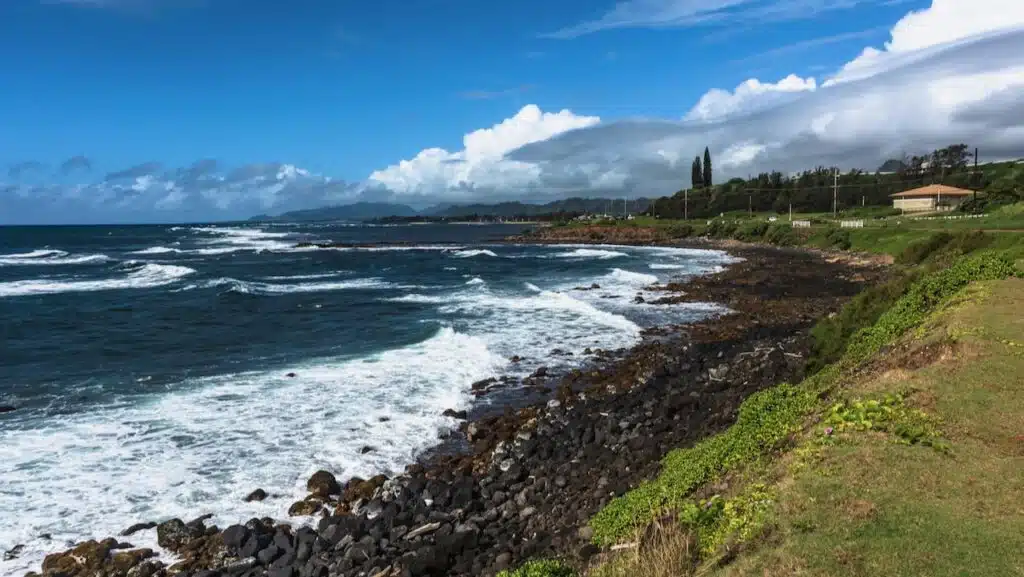View of rocky beach and white-capped waves at Kapaa on Kauai, Hawaii. A yellow house is on the right in the background.