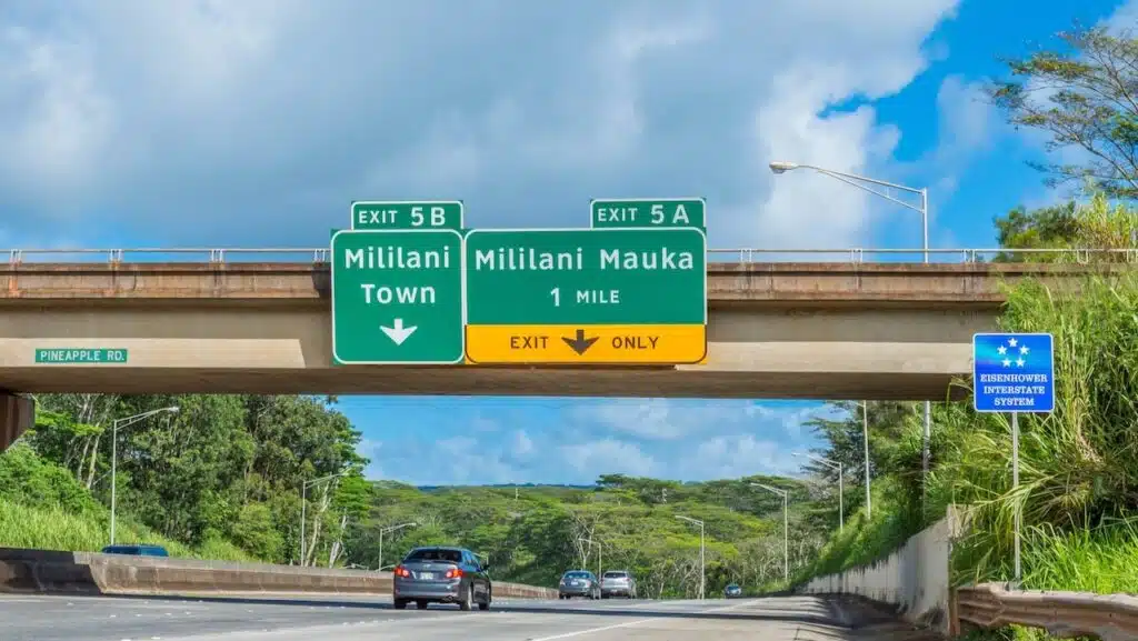 View of interstate highway on Oahu, Hawaii, with signs saying Exit 5B Militani Town with an arrow pointing down and Exit 5A Mililani Mauna 1 Mile (Exit Only in yellow with black arrow pointing down). A bridge above the highway is named Pineapple Road.