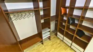 A small clothes closet with hangers on a rod and brown-wood shelves for clothes and accessories
