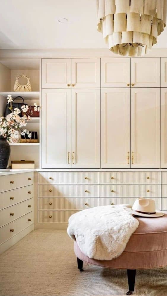 Walk-in closet with off-white cabinets and drawers and a pink ottoman in the middle.