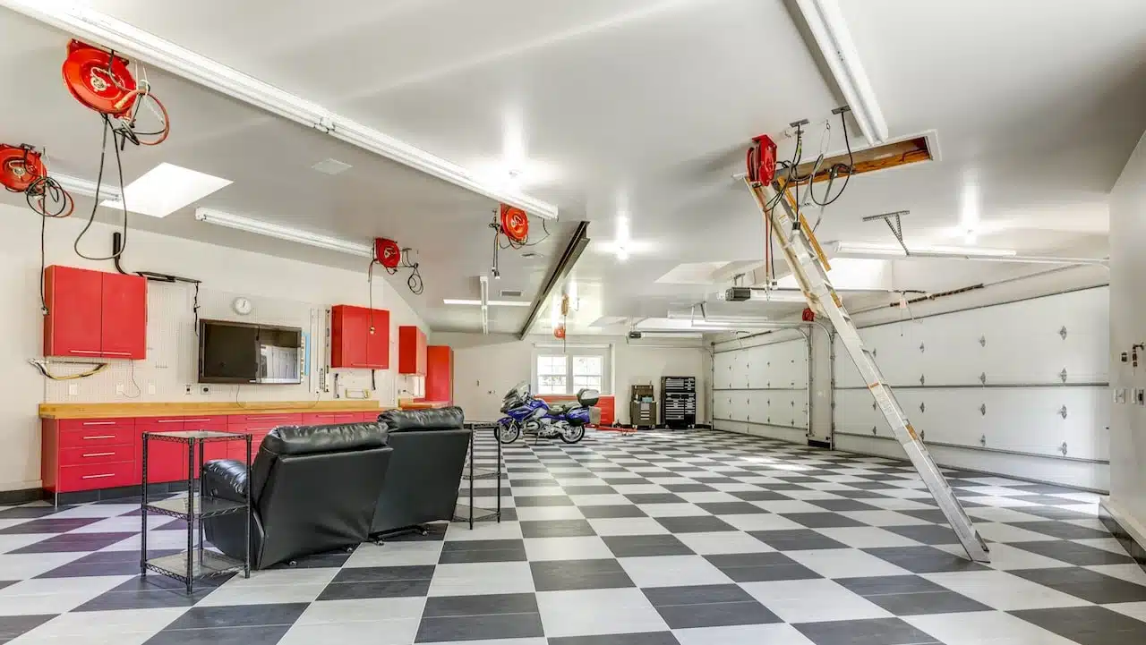 A home garage converted into a man cave with black and white checkerboard flooring, 2 black recliners and side tables with red storage and large TV along the wall. A blue motorcycle in the background along with a pulley system on the ceiling.
