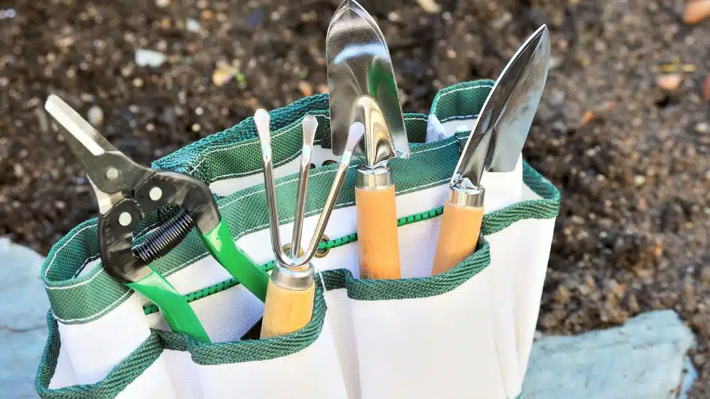 White and green canvas bag holding pruning shears, two small shoves, and a small rake.