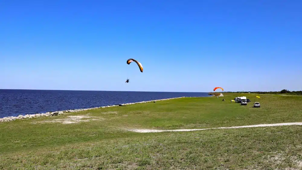 Someone paragliding over water, with other people watching from the grassy shore.