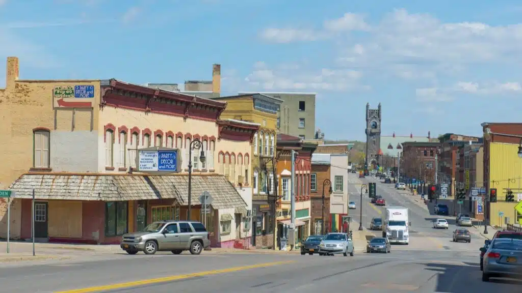 View of Main Street in Malone, New York, with sandstone and brick commercial buildings and a gray brick church bell tower in the background