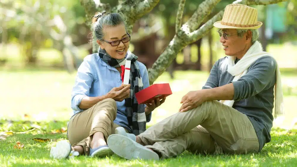 Two older people sitting on the grass in a park, looking at a gift.