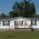 single wide modular home sits on a grassy lot surrounded by trees and blue sky