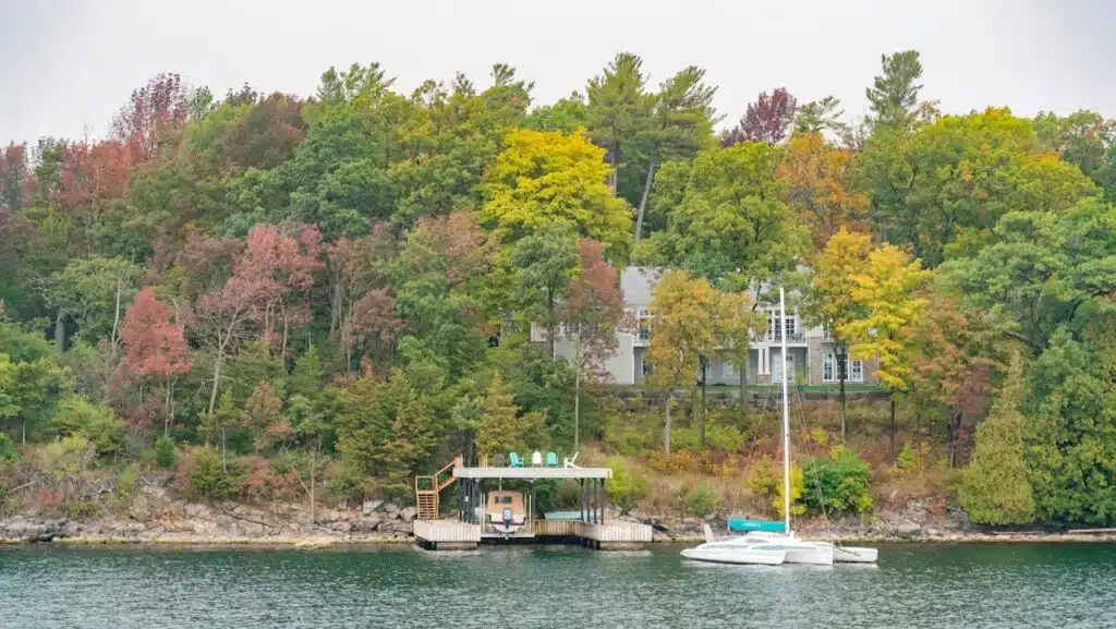 View of a home and boat dock, with a sail boat on the water, from the St. Lawrence River with autumn tree colors