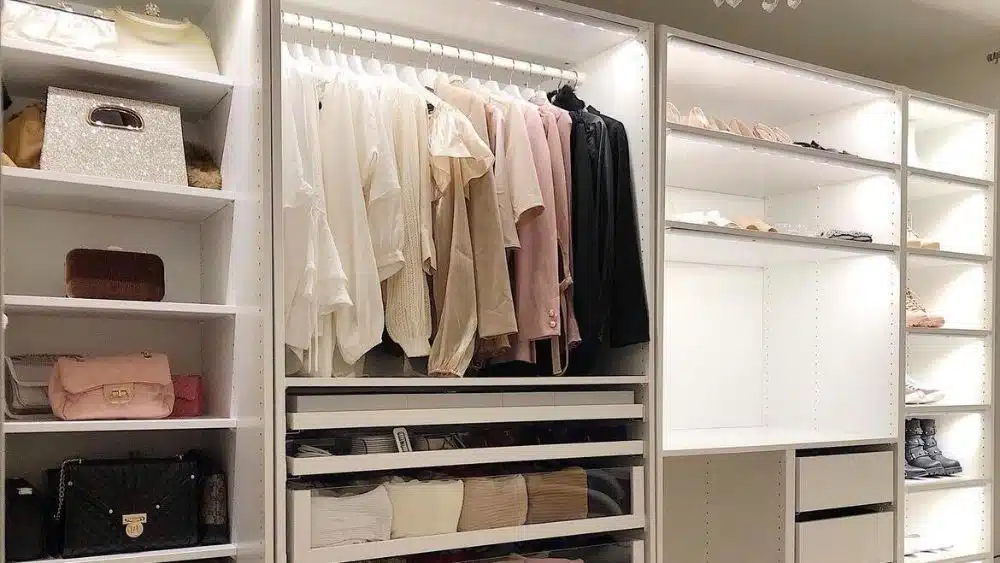 Modern closet with neutral colored clothes and open shelves that have lighting inside them.