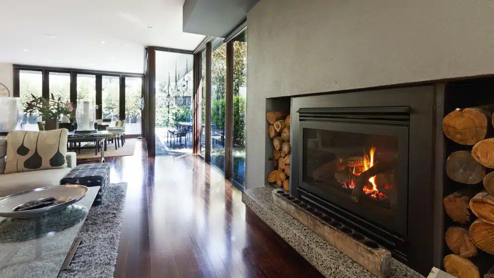 A luxury home with a large living room and a metal-framed burning fireplace with logs stacked on either side.