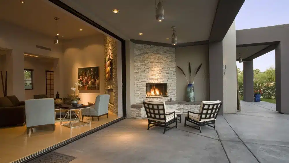 A patio next to an open-air living room. A stone fireplace is burning in the corner of the patio.