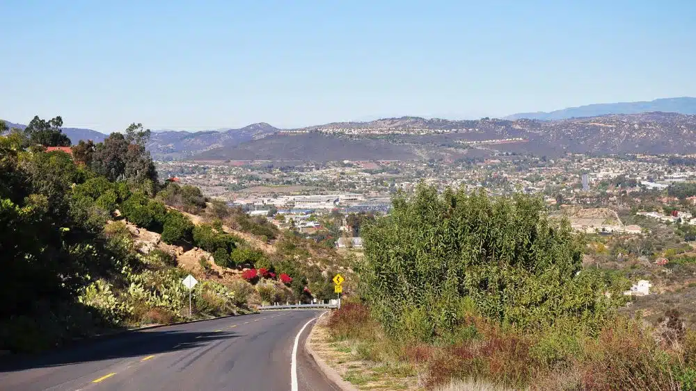 View from the side of the road in a desert, mountain area. The road is bordered by a hill covered in plants. In the distance is a city.