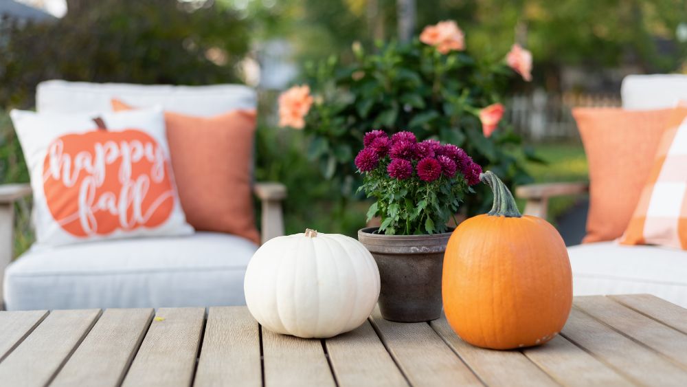 Outdoor table with two pumpkins and flowers, with seating with fall-themed pillows in the background